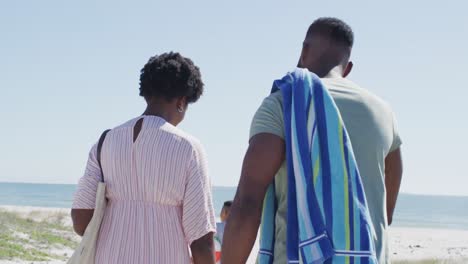 Glückliche-Afroamerikanische-Familie-Mit-Strandausrüstung,-Die-An-Einem-Sonnigen-Tag-Am-Strand-Spaziert