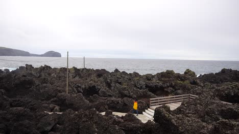 joven en chaqueta amarilla caminando en un camino entre rocas volcánicas en las piscinas naturales de varadouro, faial