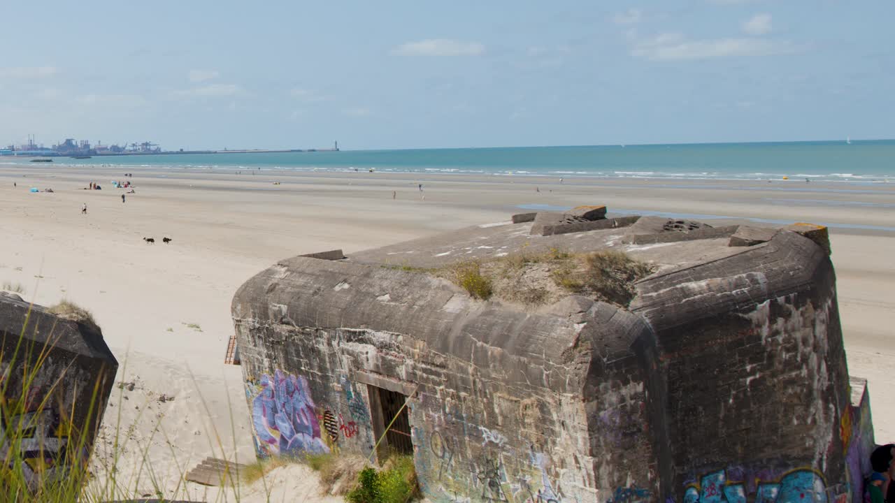 Daytime camera pan reveals graffiti-covered bunker, sandy beach, distant ocean, and scattered visitors