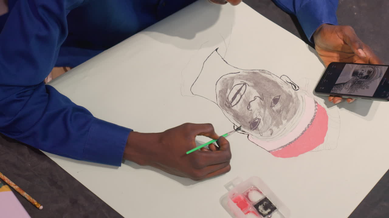 Black artist finishing black portrait painting, angled overhead of black man in blue shirt applying final fine strokes with green brush while referencing smartphone image, visible pink underlayer