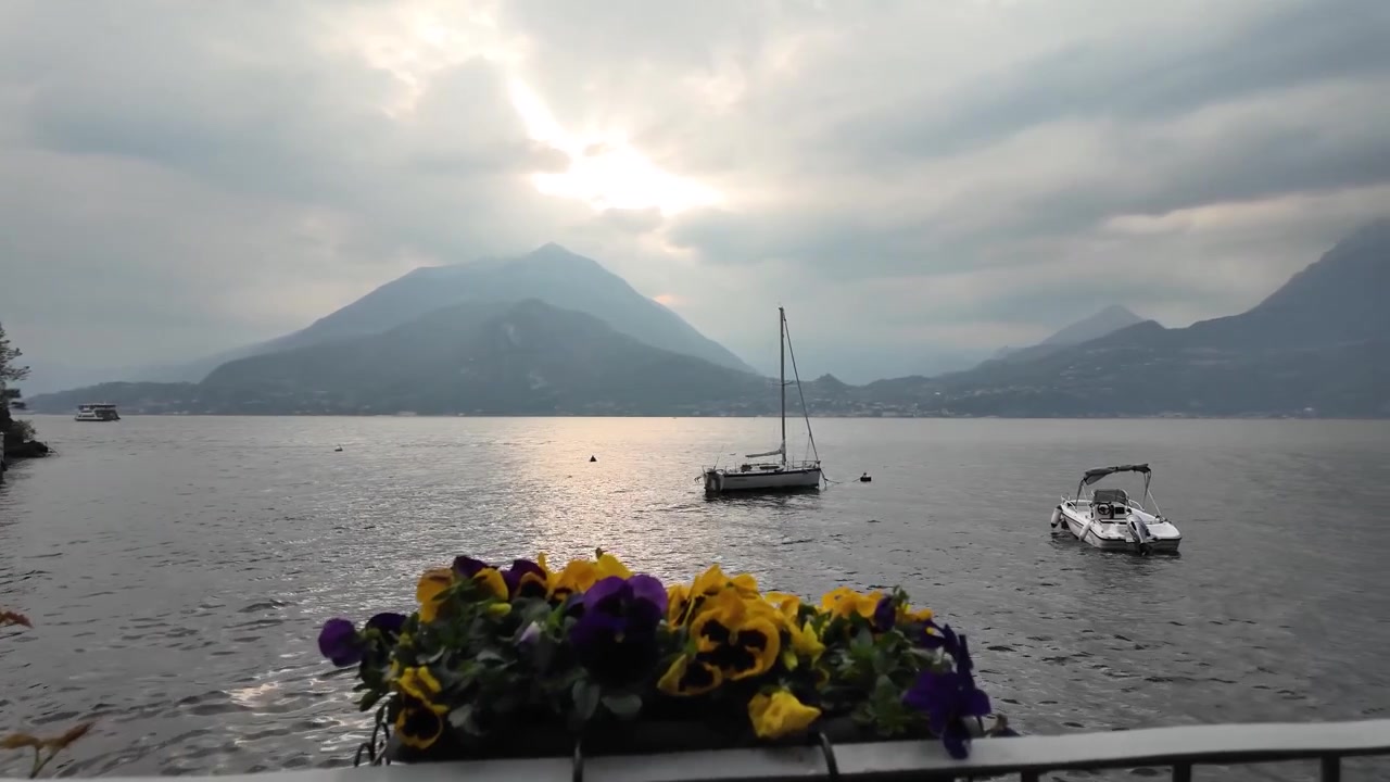Vista tranquilla del tramonto sul Lago di Como da Varenna, Italia, con barche sull'acqua, drammatiche sagome montane e fiori in primo piano.