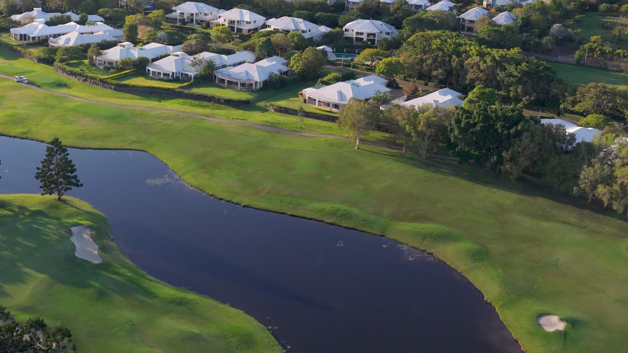 Drone footage captures a serene golf course with lush greenery, water hazards, and nearby residential areas during golden sunset hour