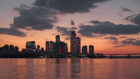 yeongdeungpo-gu skyscrapers in yeouido island silhouetted on orange sunset sky background, hangang riverbank, buildings are reflected in calm river water, traffic on mapo bridge in seoul, south korea