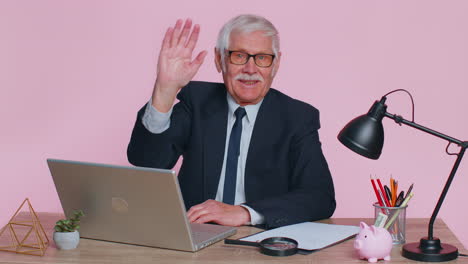 Senior-business-man-waves-hand-palm-in-hi-gesture-greeting-welcomes-someone-webinar-at-pink-office