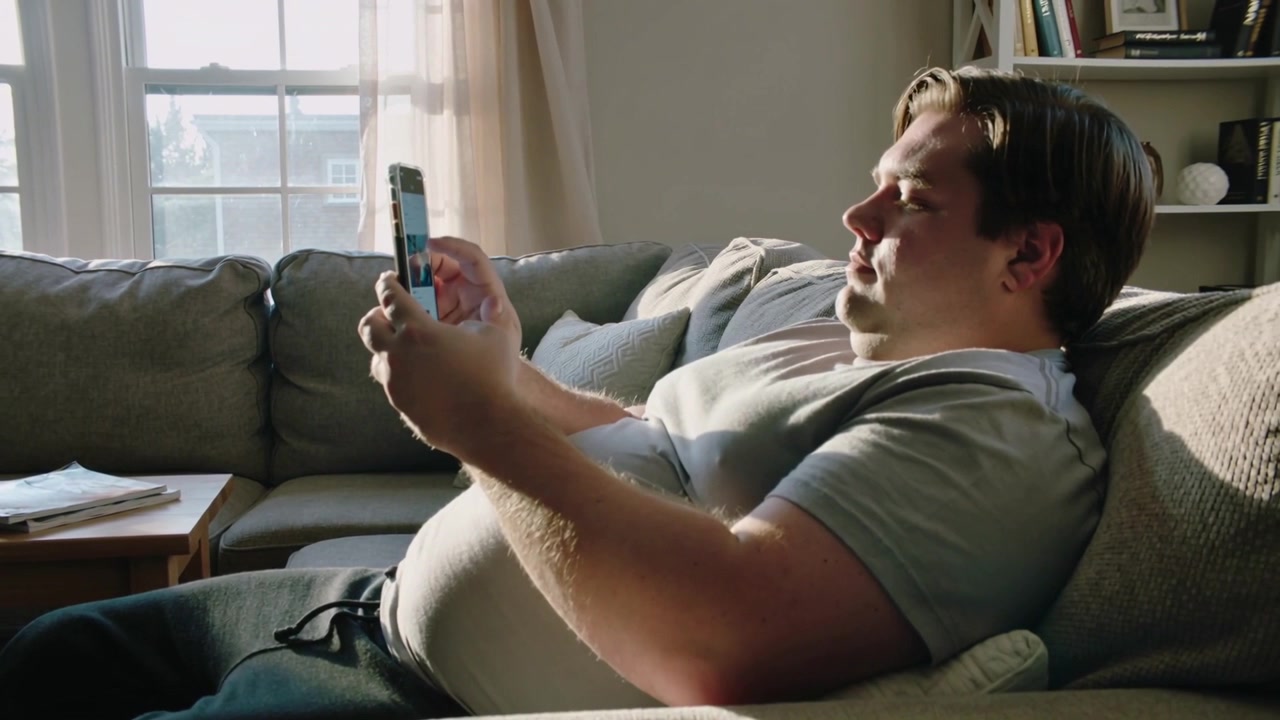 Reclining man in polo and sweatpants, tapping and scrolling smartphone on sofa at home to relax
