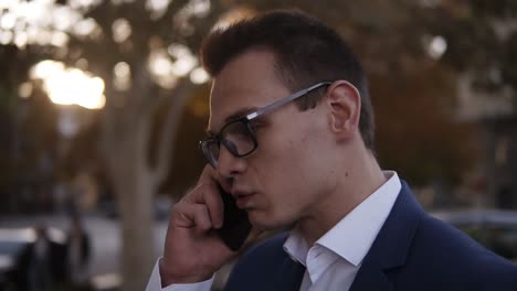 young modern businessman in stylish glasses standing on street and arguing with somebody over smartphone. blurred background