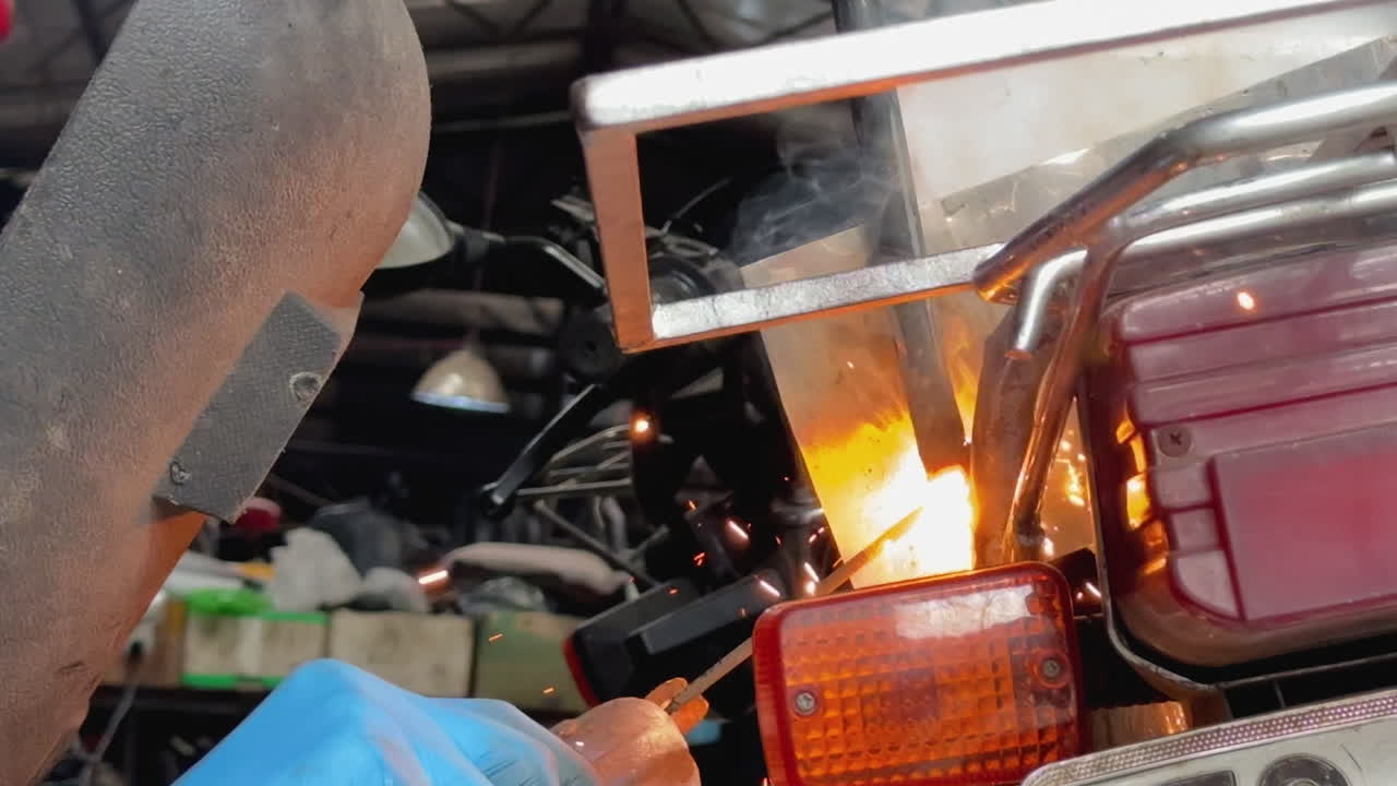 Close-up of welding sparks near a motorcycle taillight and protective mask in a metal workshop.