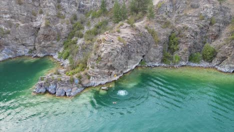 Aerial-view-of-people-jumping-off-cliff-at-rattlesnake-point-into-turquoise-water-of-Kalamalka-lake