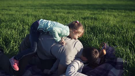 wide meadow with green grass. young father and two daughters are having fun together. fool around. the youngest has jumped on the fathers back. playing on the ground, on a picnic blanket