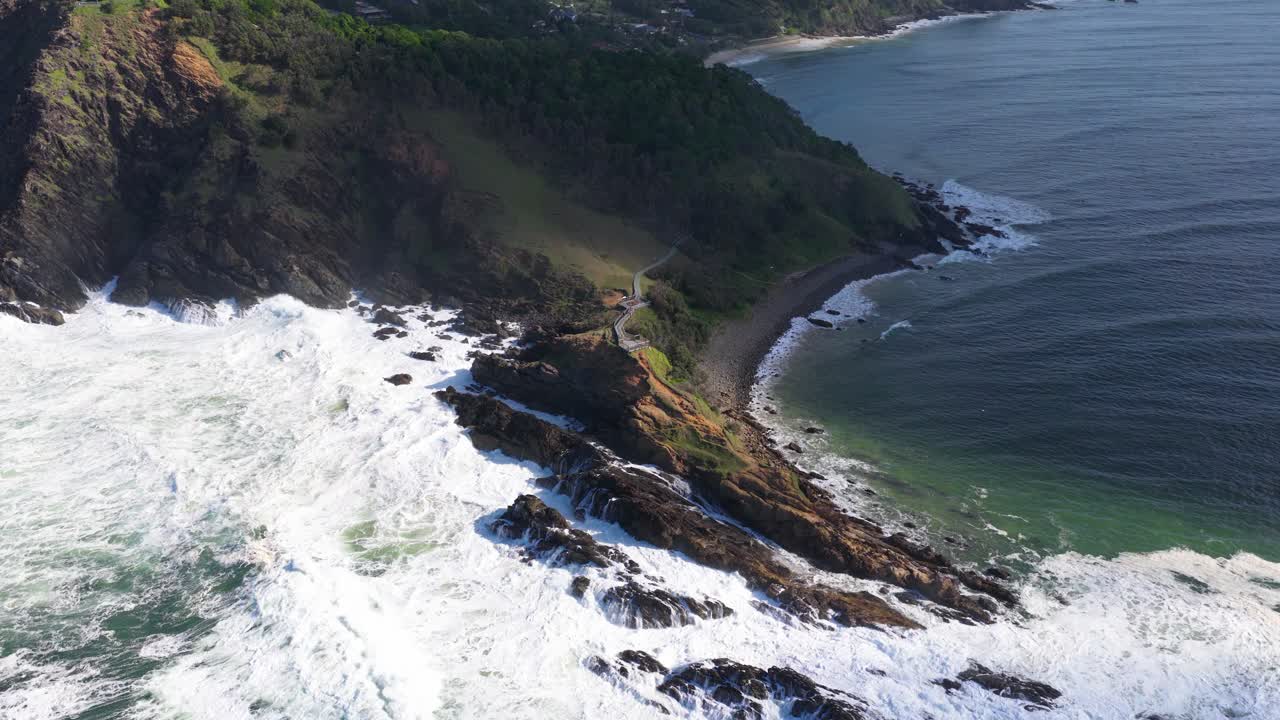 Aerial footage captures powerful ocean waves crashing against rocky cliffs at Byron Bay, Australia, under bright daylight