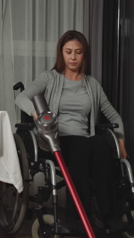 woman with muscular weakness cleans kitchen calmly using cordless vacuum cleaner. young female sits in wheelchair near kitchen table with bouquet