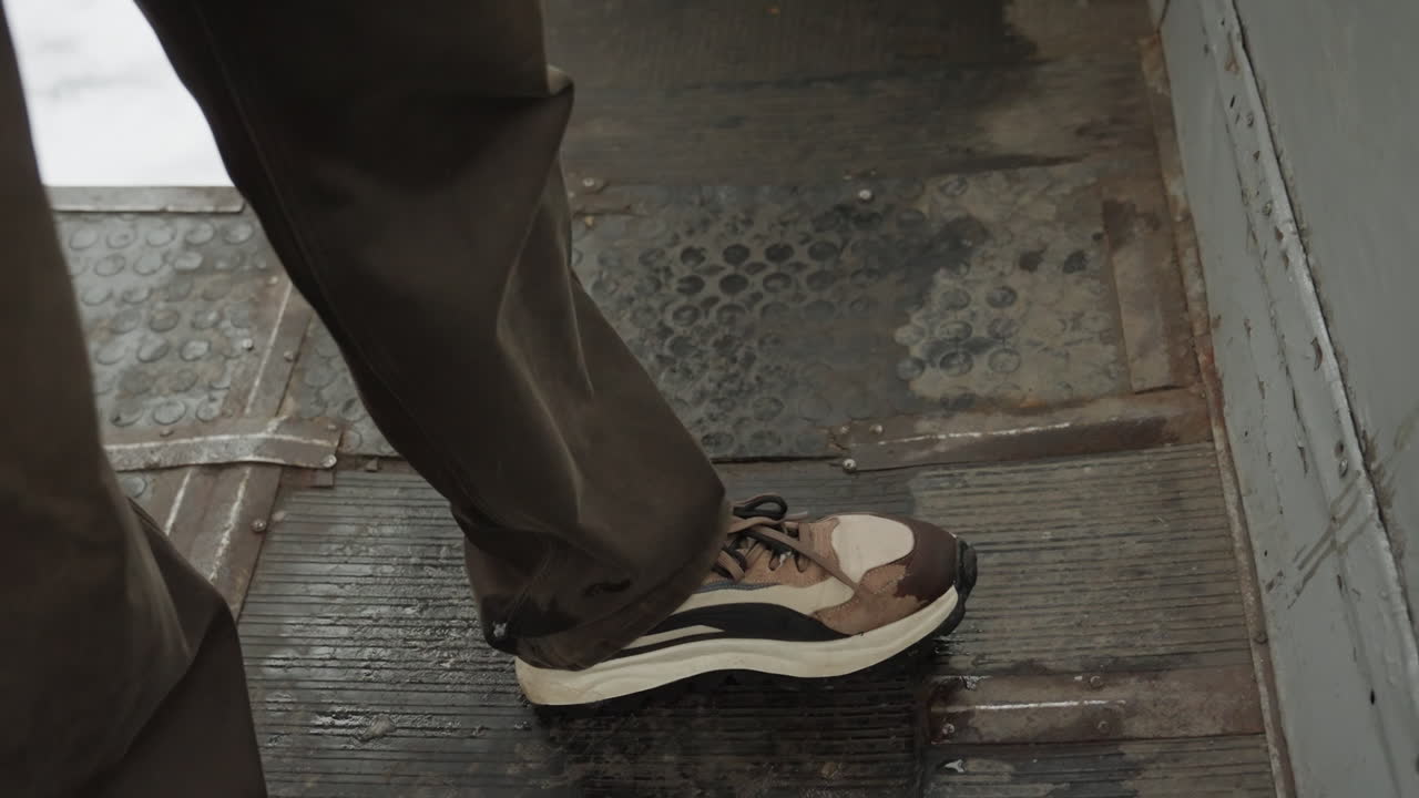 Nærbillede af sko på våde metaltrin, der viser slidt sneakersål, brune bukser træder på industriel afsats med snavs og slidt slidbane, forsigtig bevægelse og subtil balancejustering, loopbar broll-klip.