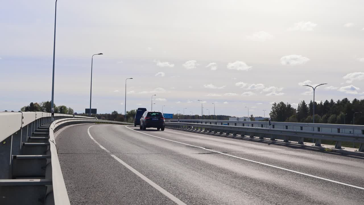 Cars driving on a bridge or an overpass during a sunny day in Laagri Estonia. The bridge goes over Pärnu highway. Sun is shining and some clouds are in the sky. Road has white markings and lamp posts.
