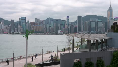 people are seen walking through victoria harbour waterfront as they enjoy their evening and skyline view of hong kong island skyscrapers in the background