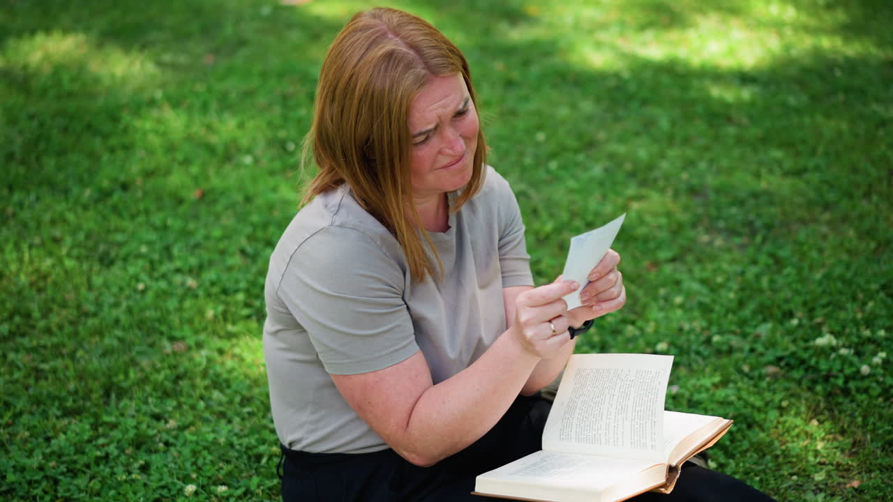 Young woman sitting on grass in quiet park flipping through book discovering hidden picture, becoming emotional and crying softly surrounded by sunlight and green nature