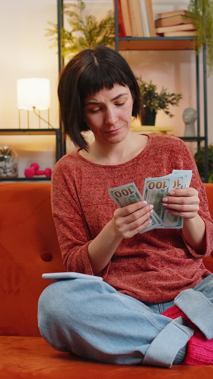 Woman counting money dollar banknotes savings earnings using smartphone calculator on sofa at home