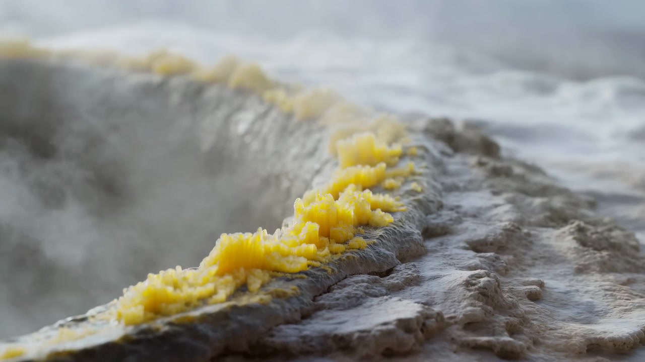 Vista dal basso del bordo di una terrazza di travertino che mostra depositi minerali giallo crema che formano una cresta frastagliata, simile a un cavolfiore, lungo il bordo di pietra stratificata bagnata vicino a una sorgente geotermica, con una leggera foschia di vapore.