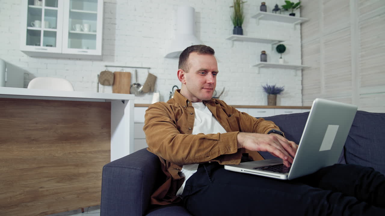 Young man working at home. Relaxed man laying on a sofa and typing on a laptop. Student studying online at home during quarantine. Remote job.