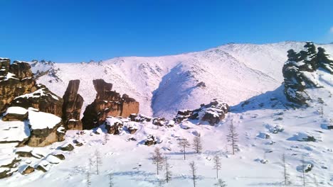 Beautiful-bird's-eye-flyover-near-winter-rocks-against-the-backdrop-of-snow-capped-mountains-and-sunny-blue-sky-4k