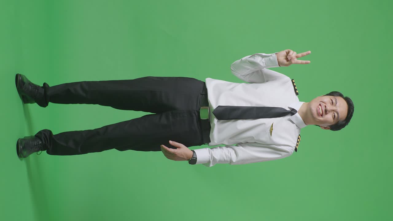 아시아 남성 조종사가 미소 짓고 녹색 화면 배경 스튜디오에 서있는 동안 카메라에 평화의 몸짓을 보여줍니다.
