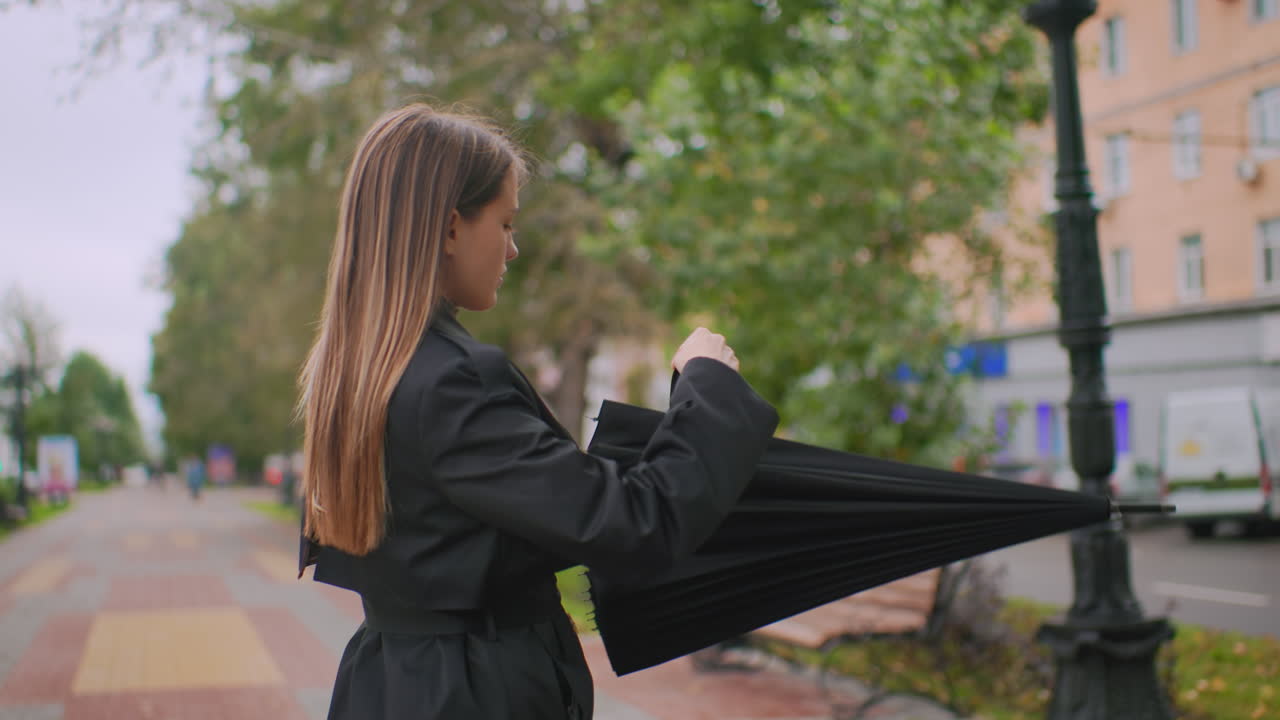 Beautiful girl in black coat, closing her black umbrella on city sidewalk with trees, benches, cars, and buildings in background during cloudy autumn day,calm, casual, peaceful, leisure