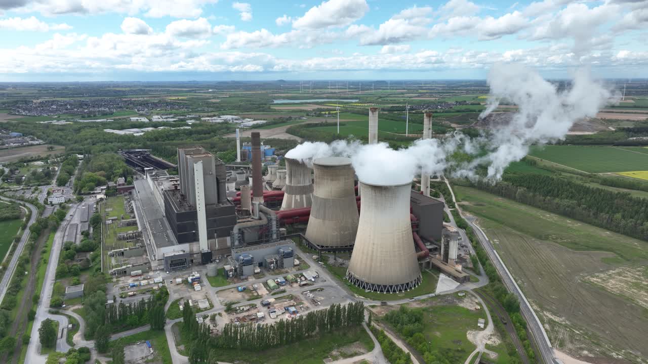 Aerial view of a large power plant with cooling towers emitting steam, surrounded by green fields and distant wind turbines