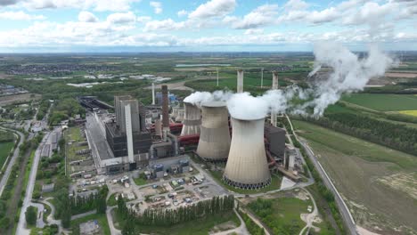 aerial view of a large power plant with cooling towers emitting steam, surrounded by green fields and distant wind turbines