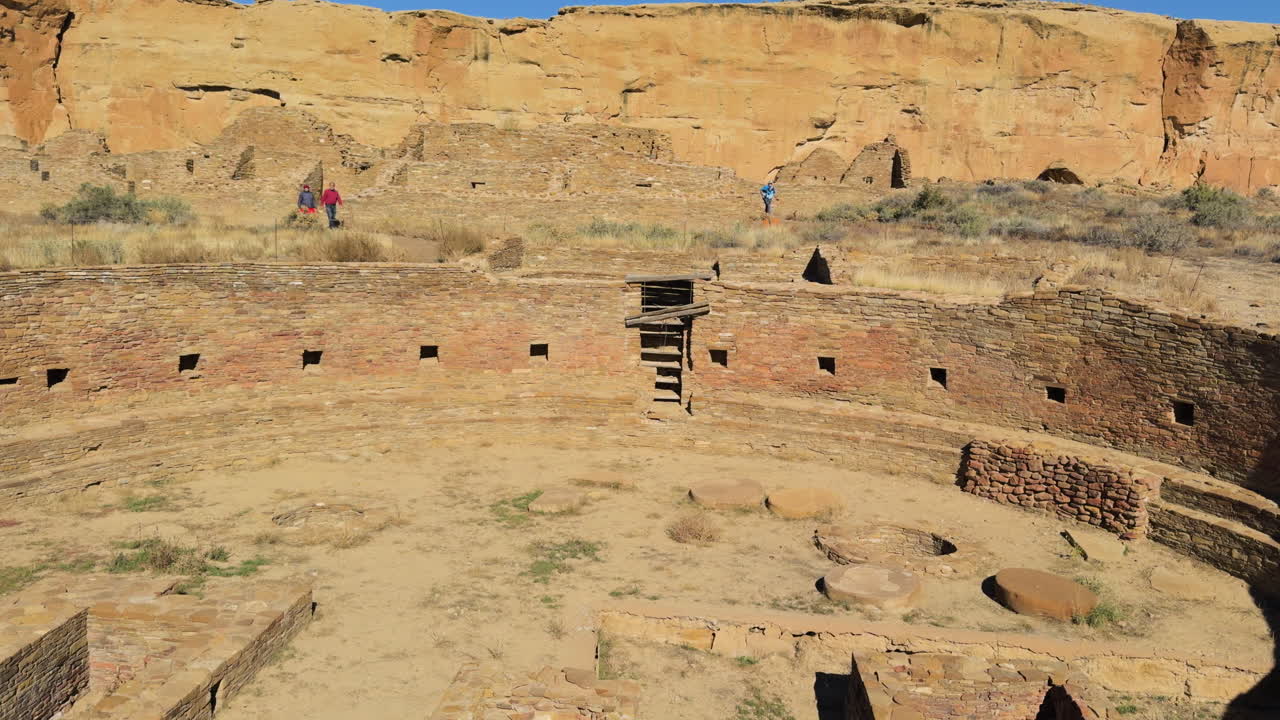 Wide view of circular kiva ruins where foreign visitors walking around in desert terrain at Chaco Canyon, New Mexico.