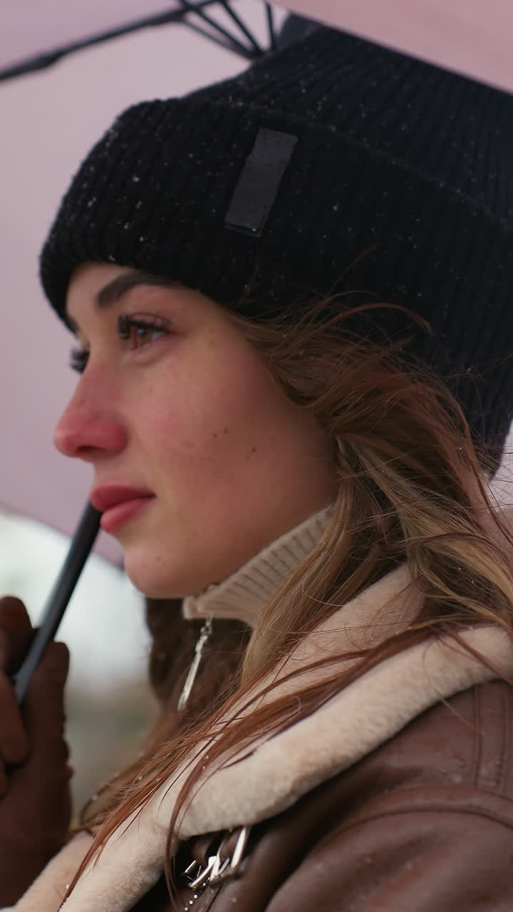 Cheerful young woman in black knit cap and brown shearling jacket holding umbrella while walking slowly on cold overcast day, snowflakes on her hat, enjoying quiet moment outdoors in urban winter