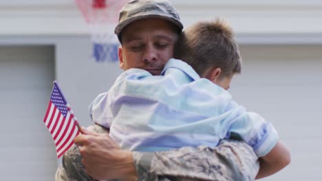 Happy-caucasian-male-soldier-hugging-his-smiling-son-holding-flag-in-garden-outside-their-house