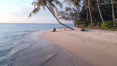 Profesora-De-Yoga-Estirándose-En-La-Playa-Bajo-La-Vista-Aérea-Mágica-De-Palmeras,-El-Auge-Del-Vuelo-Se-Desliza-Hacia-El-Dron-Derecho