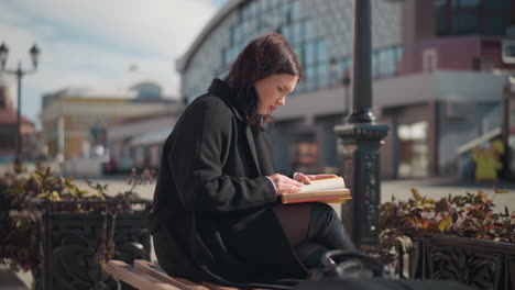 vista trasera de una señora centrada en su libro mientras está sentada al aire libre, rodeada de elementos urbanos, incluidos potes de flores y vitrinas borrosas