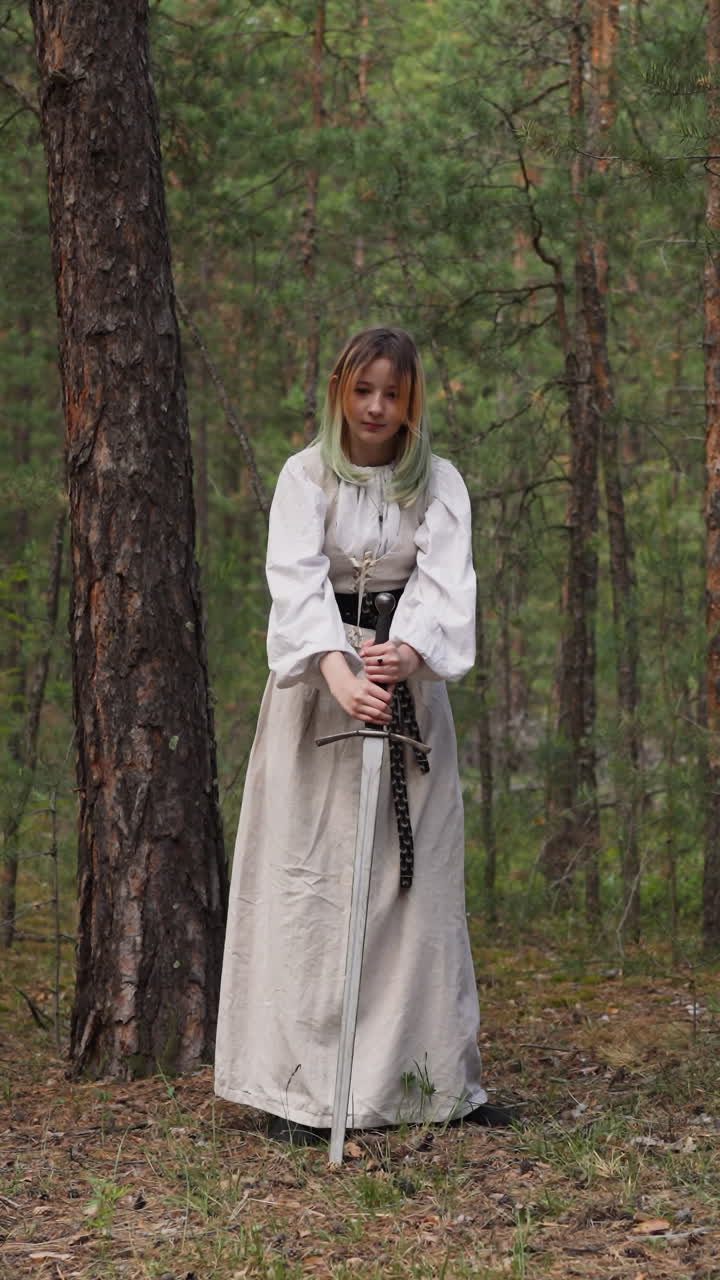 una chica adolescente bonita en un vestido medieval blanco clava una espada en el suelo en el bosque de pinos. jugador de rodillos infantil en el campamento histórico. atmosfera de cola de hadas