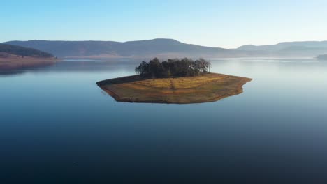 Flying-towards-a-lonely-island-surrounded-by-blue-water,-mountains-and-clear-skies-during-the-autumn