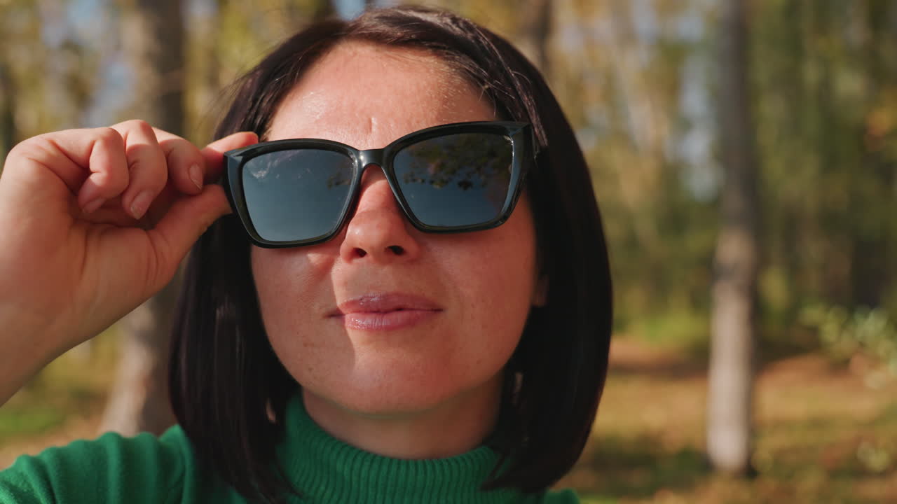 Mulher de camisola verde a ajustar os óculos de sol a acampar entre bétulas, campista relaxada a sorrir enquanto testa o reflexo da lente, luz solar a brilhar nas armações, brisa suave entre as folhas, retrato em close-up