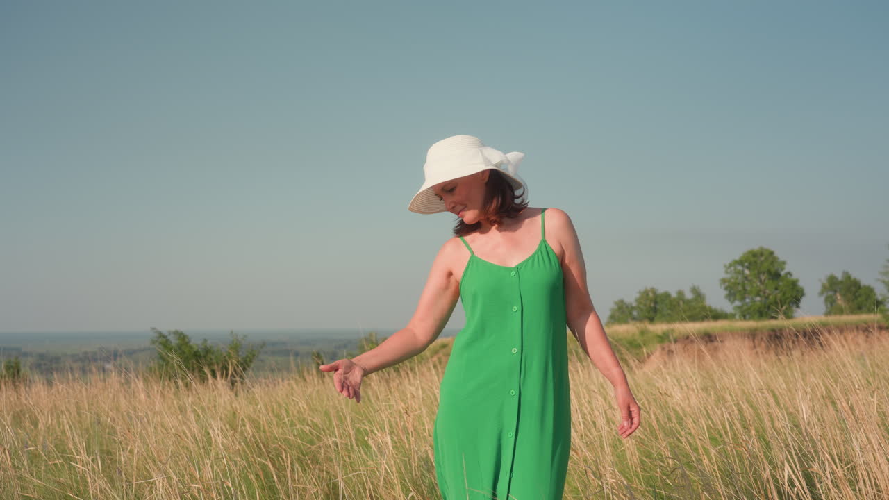 woman in green dress and white hat gently poses in golden grass meadow under clear sky, touching tall dry stalks while enjoying peaceful summer breeze, expressing calm, connection