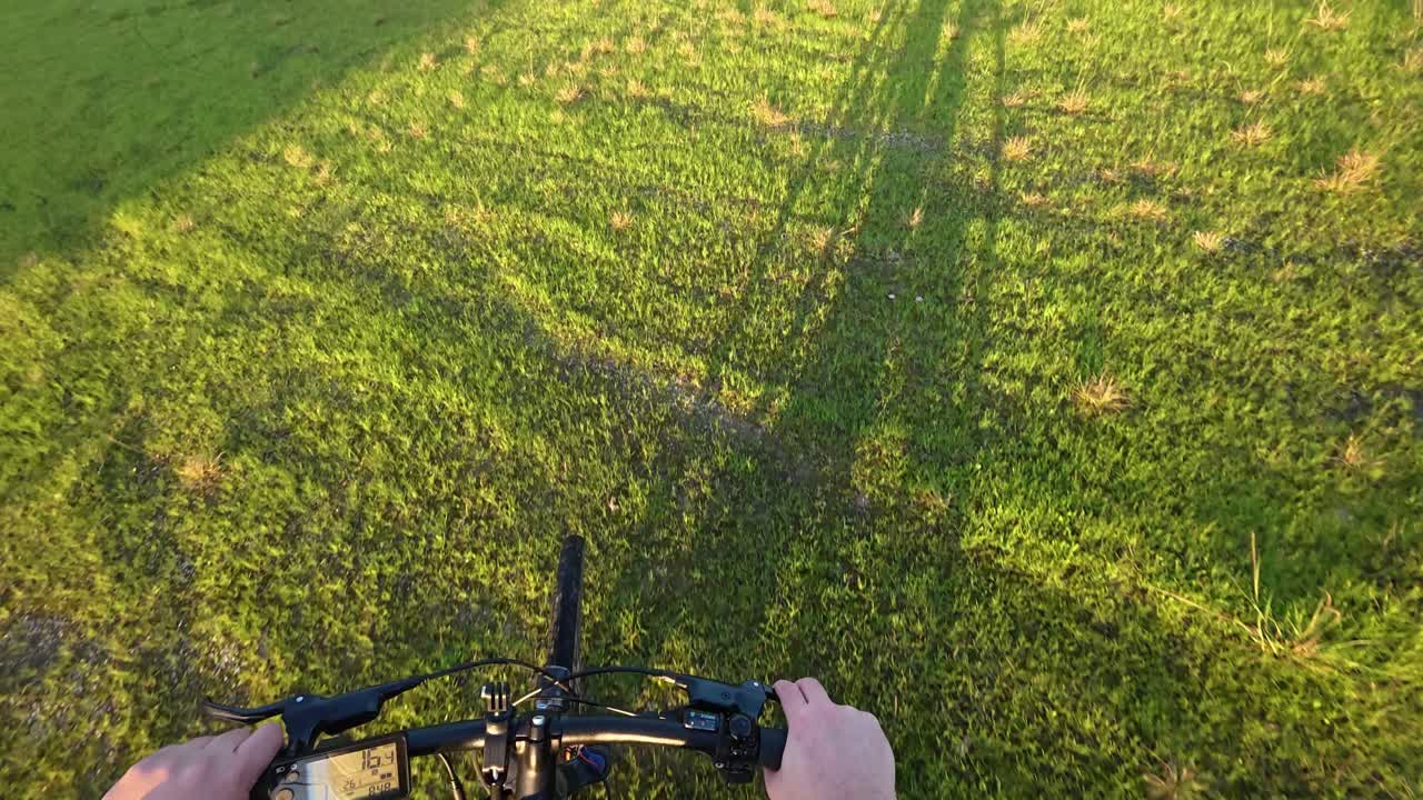 잔디을 가로질러 자전거를 타고 가는 자전거 타는 사람의 포인트 오브 뷰 으로, 그들의 그림자가 빛으로 밝은 땅에서 보인다.