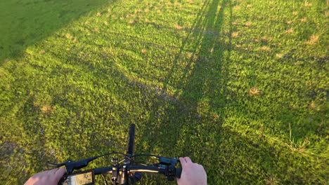 una toma de punto de vista de un ciclista montando a través de un campo cubierto de hierba, con su sombra visible en el suelo iluminado por el sol.