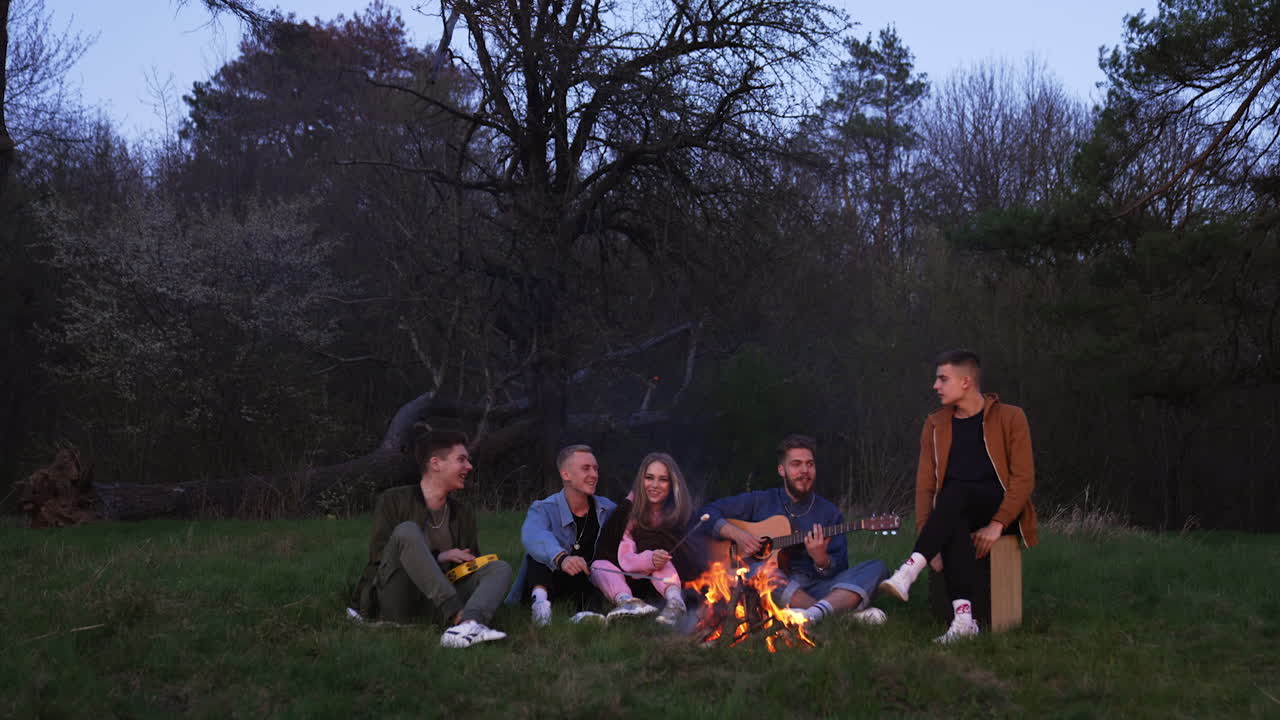 Close friends sitting on the grass in front of campfire. Couple in the middle frying marshmallows on the fire. Guys creating good mood by playing musical instruments.