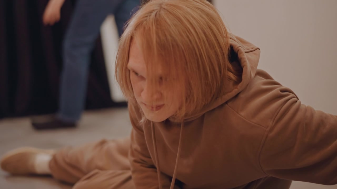 White student rehearsing floor choreography in studio, ginger hair and brown hoodie, focused expression, coach offframe offering guidance, slowmotion floor transitions on parquet, dim curtains