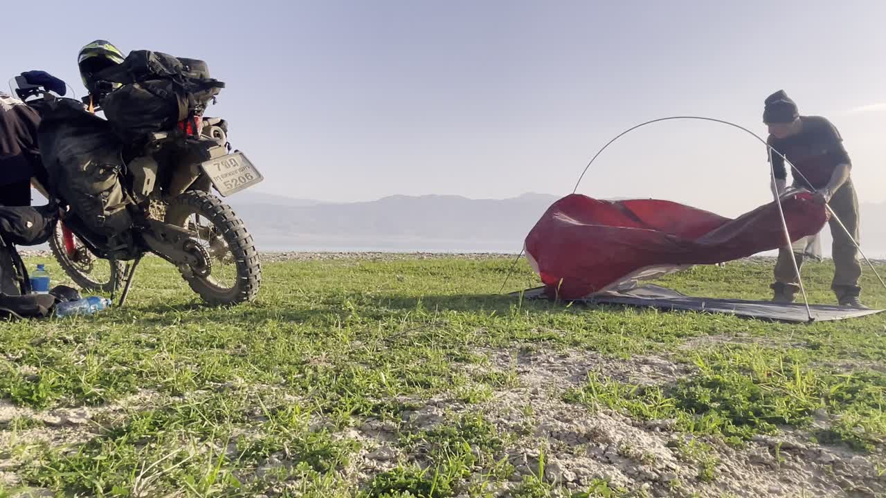 Viaggiatore avventuroso che monta una tenda con una moto da turismo completamente carica parcheggiata sull'erba, sullo sfondo di montagne.