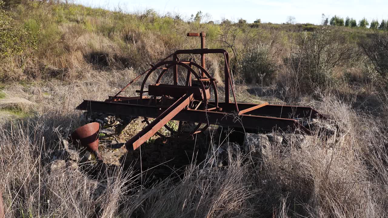 Primo piano di un antico motore agricolo, ricoperto di ruggine, costruito per estrarre acqua da un pozzo profondo e per sostenere i raccolti durante innumerevoli stagioni secche, circondato da erba dorata secca.