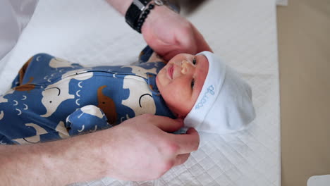 little caucasian newborn in a blue romper is put a white cap on. dad's hands dressing a baby lying on the sheet. close up.