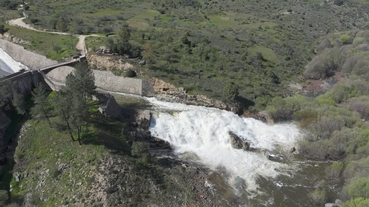 Drone footage of powerful dam discharge forming a spectacular white waterfall splitting into two, shaped like a horse tail due to the force of the water, in a natural setting