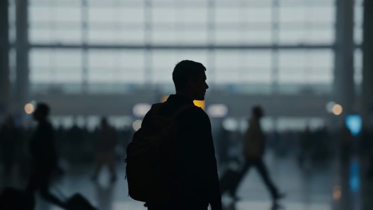 Central silhouetted traveler stands with backpack facing expansive arched glass facade inside modern airport concourse while busy passengers, wheeled luggage, reflections, and deep interior