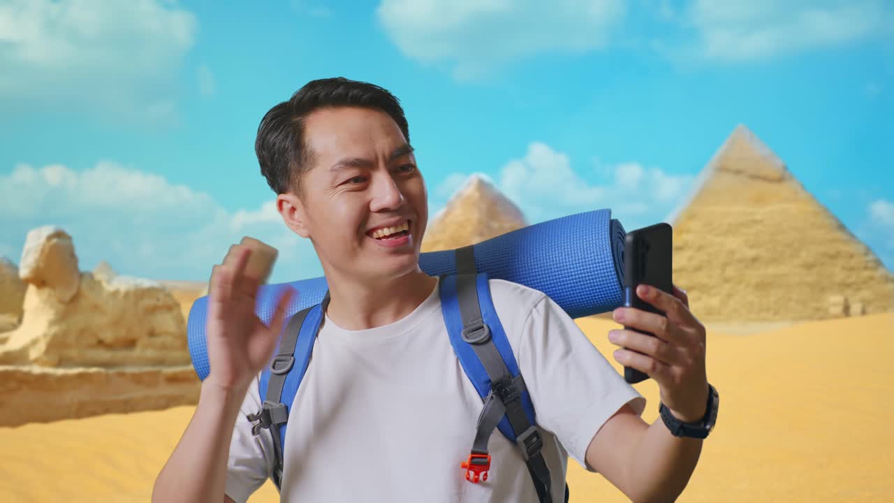 Close Up Of Asian Male Hiker With Mountaineering Backpack Waving His Hand And Pointing Back While Having A Video Call On Smartphone While Traveling In Pyramid Of Giza