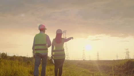 The-view-from-the-back:-group-of-engineers-at-a-high-voltage-power-plant-with-a-tablet-and-drawings-walk-and-discuss-a-plan-for-the-supply-of-electricity-to-the-city.-Transportation-of-renewable-clean-energy.