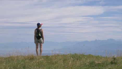 A-lone-female-hiker-stands-on-a-mountain-top-looking-at-the-view-of-a-large-alpine-lake-and-surrounding-alpine-peaks