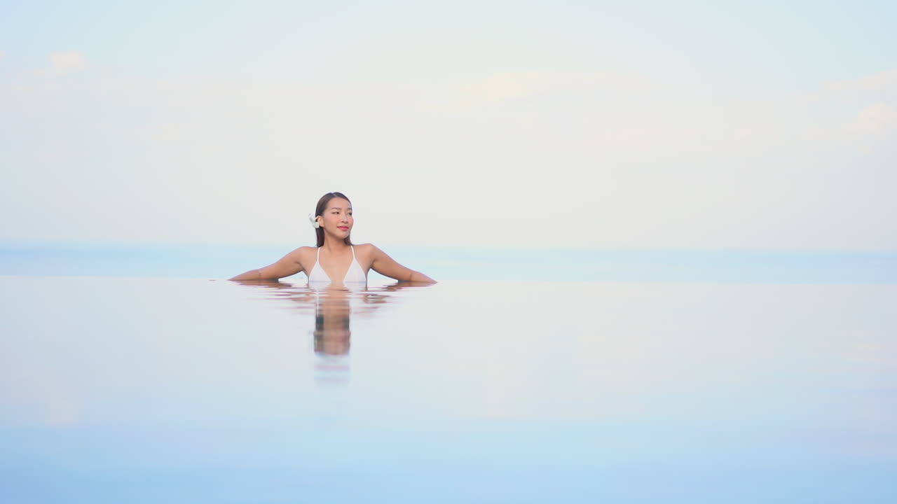 Attraktive asiatische Frau mit Blume hinter dem Ohr, die sich in einem Infinity-Pool eines Resorts entspannt, mit einer Spiegelung auf der Wasseroberfläche.