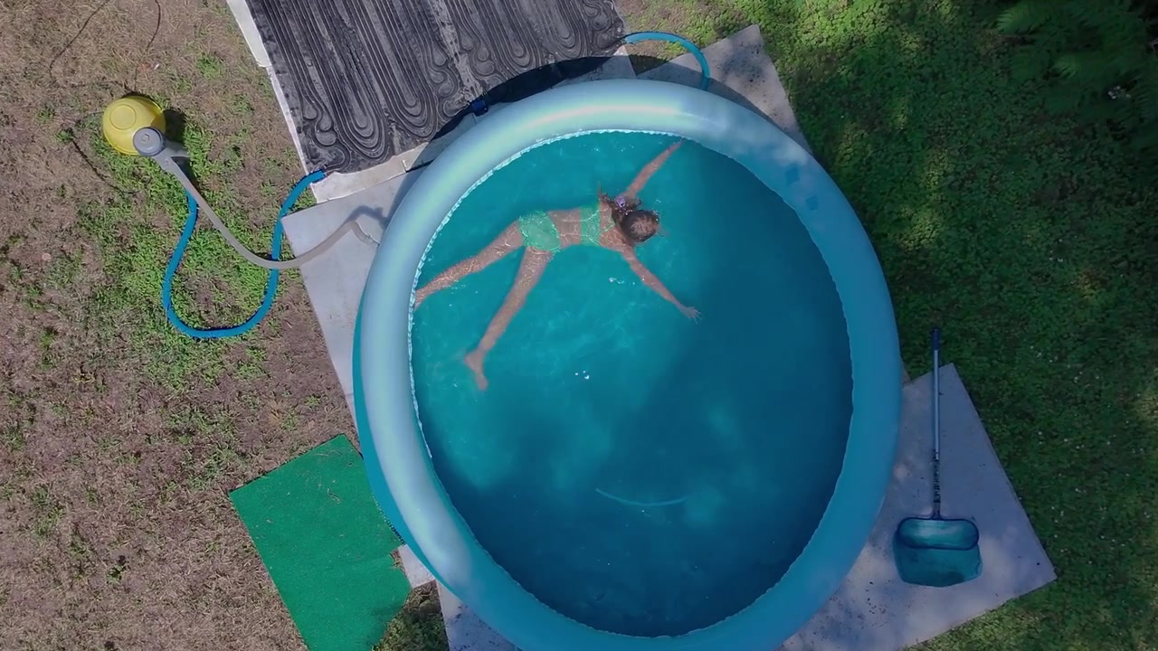Luftfoto fra høj vinkel, der zoomer fra en bred udsigt over gården til et nærbillede af en pige i før-teenagealderen, der svømmer i det klare blå vand i en rund oppustelig pool i en solrig grøn baghave